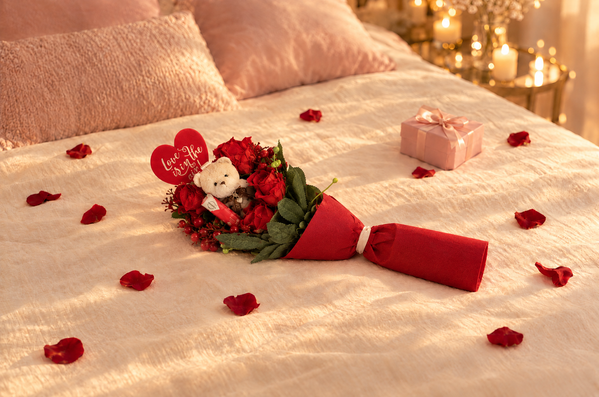 Falling in Love bouquet lying on a cream bedspread with rose petals and candlelight